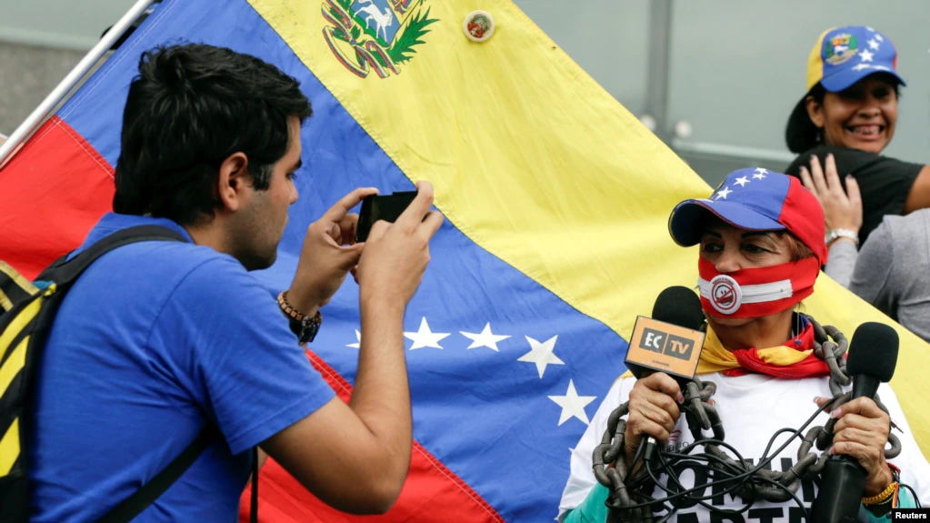 ARCHIVO - Un hombre toma una fotografía de una mujer que, mientras sostiene micrófonos, lleva una mordaza y una cadena. Fue durante una manifestación para conmemorar el día Mundial de la Libertad de Prensa en Caracas, Venezuela, el 3 de mayo de 2016