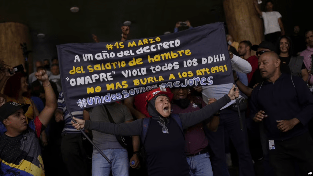 Un manifestante sostiene un cartel que exige mejores salarios mientras los partidarios del gobierno se paran detrás en un intento de que los manifestantes abandonen el Ministerio de Trabajo en Caracas, Venezuela, el miércoles 15 de marzo de 2023. (Foto AP/Matías Delacroix)